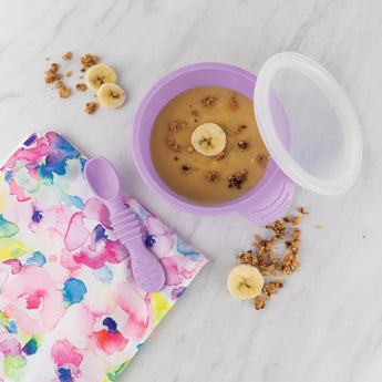 Bumkins Silicone First Feeding Set: Lavender with applesauce surrounded by banana slices, granola, and a floral pouch on a marble surface.
