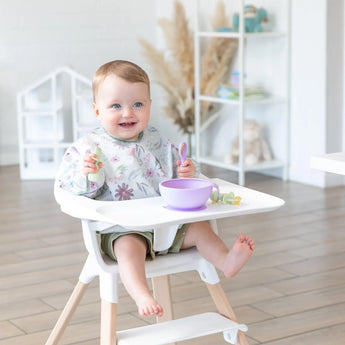 A baby in a high chair wearing a floral bib eats from a Bumkins Silicone First Feeding Set: Lavender with green utensils in a bright room.