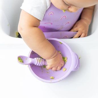 A baby in a high chair eats mashed avocado from a Bumkins Silicone First Feeding Set: Lavender while wearing a matching silicone bib.