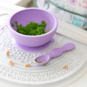 Close-up of Bumkins Silicone First Feeding Set: Lavender filled with green mashed food on a white silicone mat adorned with food crumbs.