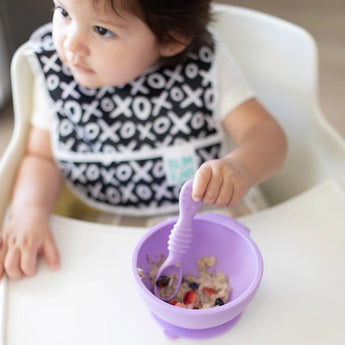 A child in a high chair with a bib enjoys oatmeal and berries from a Bumkins Silicone First Feeding Set: Lavender