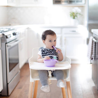 A baby in a high chair uses a Bumkins Silicone Dipping Spoons 3 Pack: Lollipop to eat from a lavender bowl in a bright, modern, kitchen.