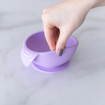 A hand holds a Bumkins Silicone First Feeding Set: Lavender, demonstrating the suction cup bottom on a marble surface.