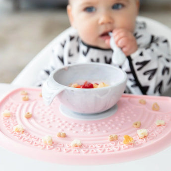 A baby in a black & white bib eats from a Bumkins Silicone First Feeding Set: Marble on a pink silicone mat covered in cereal pieces.