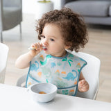 A curly-haired toddler in dinosaur bib eats from a Bumkins Silicone First Feeding Set: Marble at a white table.