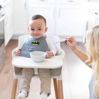 A baby in a high chair, wearing a gray bib, is fed by an adult with a Bumkins Silicone Dipping Spoons 3 Pack: Taffy from a gray bowl.