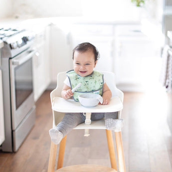 A baby in a high chair smiles, wearing green bib in a bright kitchen, eating from a bowl with a Bumkins Silicone Dipping Spoons 3 Pack: Taffy.