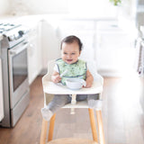 A happy baby in a high chair smiles in a bright kitchen, ready to eat from a Bumkins Silicone First Feeding Set: Marble.