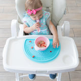 A toddler in a high chair with a pink bow enjoys cereal and strawberries from a Bumkins Silicone First Feeding Set: Pink on a blue placemat.