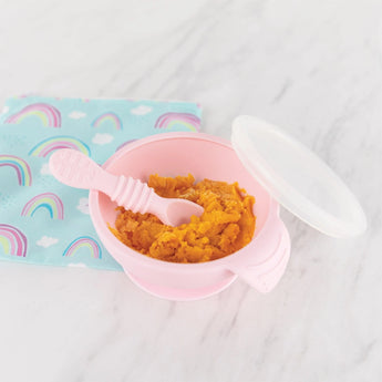 Bumkins Silicone First Feeding Set: Pink with orange mash on a marble surface next to a rainbow print pouch.