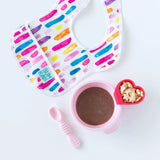 A colorful bib sits next to Bumkins Silicone First Feeding Set: Pink with puree and a red heart-shaped cup of cereal on a white background.