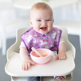 A baby in a purple unicorn-print bib sits in a high chair with Bumkins Silicone First Feeding Set: Pink in a bright room.
