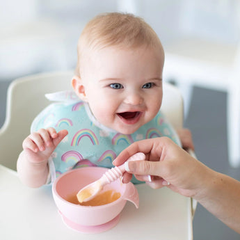 A smiling baby sits in a high chair as an adult feeds them from a pink bowl with a pink Bumkins Silicone Dipping Spoons 3 Pack: Lollipop.