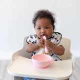 A baby in a high chair wearing a black & white bib eats from a Bumkins Silicone First Feeding Set: Pink.