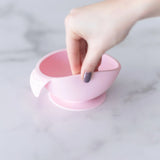 A hand holds a Bumkins Silicone First Feeding Set: Pink, demonstrating the suction cup bottom on a marble surface.