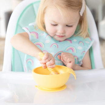 A child in a high chair wearing a rainbow-print bib uses a yellow Bumkins Silicone Dipping Spoons 3 Pack: Tutti-Frutti to eat from a bowl.
