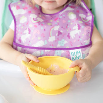 A child in a high chair in a purple bib holds a yellow bowl with a yellow Bumkins Silicone Dipping Spoons 3 Pack: Tutti-Frutti inside.