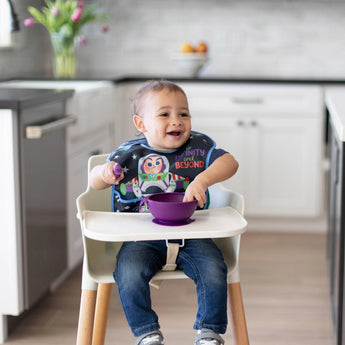 In a modern kitchen, a smiling toddler in a high chair wearing a space-themed bib holds a Bumkins Silicone First Feeding Set: Purple.