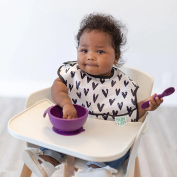 A baby in a high chair wearing a black & white bib reaches into a Bumkins Silicone First Feeding Set: Purple.