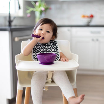 A toddler in a high chair enjoys a meal from a Bumkins Silicone First Feeding Set: Purple in a bright kitchen.