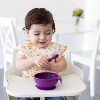 A toddler with brown hair in a high chair uses a Bumkins Silicone First Feeding Set: Purple while wearing a bib with yellow patterns.