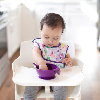In a bright kitchen, a toddler in a high chair  wearing a colorful bib uses a Bumkins Silicone First Feeding Set: Purple.