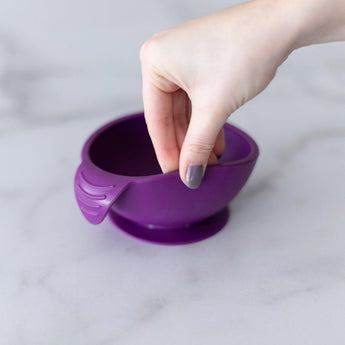 A hand holds a Bumkins Silicone First Feeding Set: Purple, demonstrating the suction cup bottom on a marble surface.