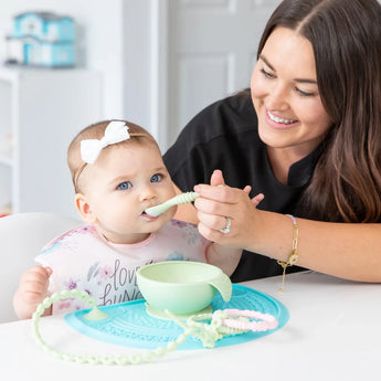 A woman feeds a baby in a floral bib from a Bumkins Silicone First Feeding Set: Sage on a blue silicone placemat next to a teething toy.