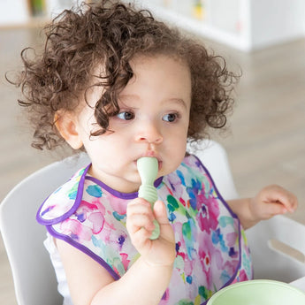 A toddler with curly hair sits at a table, holding a spoon from Bumkins Silicone First Feeding Set: Sage, while wearing a colorful bib.