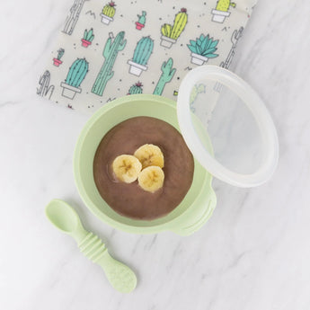 Bumkins Silicone First Feeding Set: Sage with chocolate pudding topped with bananas on a marble background next to a cactus print pouch.