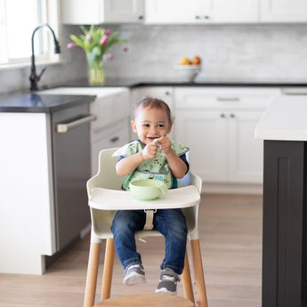 A toddler in a green bib smiles in a high chair, eating from a Bumkins Silicone First Feeding Set: Sage in a bright kitchen.