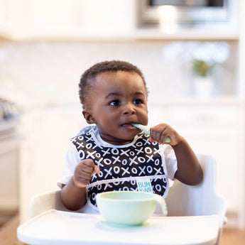 A toddler in a high chair eats from a Bumkins Silicone First Feeding Set: Sage while wearing a black & white printed bib.