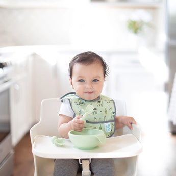 A baby in a high chair smiles in a bright kitchen, holding a baby spoon over a Bumkins Silicone First Feeding Set: Sage.