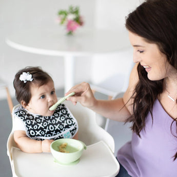 A woman smiles as she feeds a baby in a high chair from a green bowl with a Bumkins Silicone Dipping Spoons 3 Pack: Taffy.