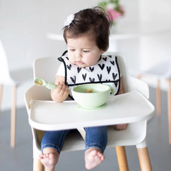 A baby in a high chair wearing a black & white bib holds a green Bumkins Silicone Dipping Spoons 3 Pack: Gumdrop near a green bowl.