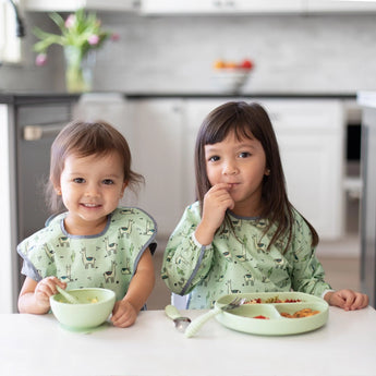 Two kids sits at a white table wearing matching green bibs with animal prints; one eats from a Bumkins Silicone First Feeding Set: Sage.