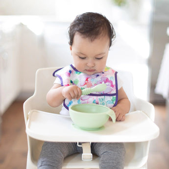 A baby sits in a high chair, holding a Bumkins Silicone Dipping Spoons 3 Pack: Taffy and curiously eyeing a green bowl on the tray.