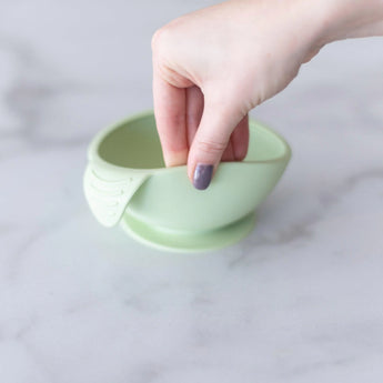 A hand holds a Bumkins Silicone First Feeding Set: Sage, demonstrating the suction cup bottom on a marble surface.