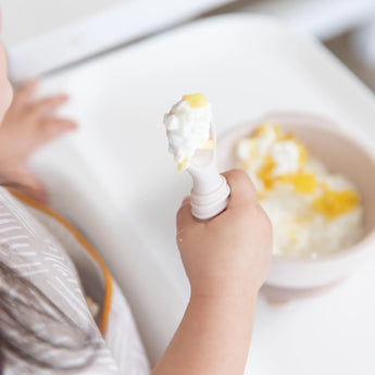 Close-up of a child holding a spoon from Bumkins Silicone First Feeding Set: Sand covered in yogurt, with the bowl blurred underneath.