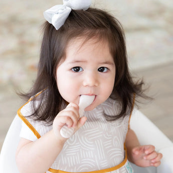 A toddler in a high chair with a white bow and bib chews a spoon from Bumkins Silicone First Feeding Set: Sand.