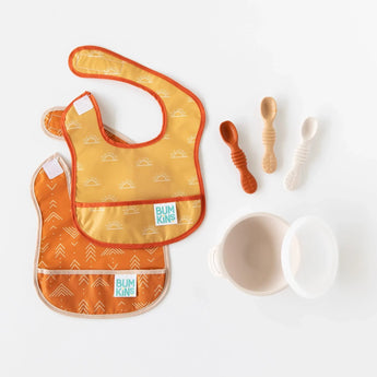 Bumkins Silicone First Feeding Set: Sand sits among baby spoons and colorful bibs on a white background.