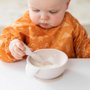 A baby joyfully eats yogurt from Bumkins Silicone First Feeding Set: Sand at a white table, wearing an orange bib.