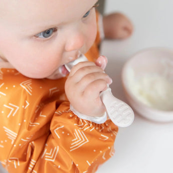 Close-up of a baby in an orange bib joyfully self-feeding yogurt from Bumkins Silicone First Feeding Set: Sand.