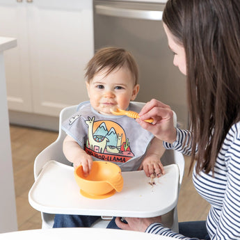 An adult uses an orange Bumkins Silicone Dipping Spoons 3 Pack: Tutti-Frutti to feed a happy baby with an orange bowl in a high chair.