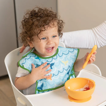A toddler beams in a high chair wearing a Bumkins SuperBib® and Sleeved Bib 3-Piece Set: Rolling With The Waves, Whale Tail, Ocean Life.