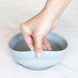 A hand holds a Bumkins Silicone Grip Bowl: Gray, demonstrating the suction cup bottom on a marble surface.
