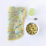 A folded camping-themed bib sits next to Bumkins Silicone Grip Bowl: Marble with pasta & peas, a fork, & a green cup on a white background.