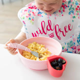 A child eats scrambled eggs from Bumkins Silicone Grip Bowl: Pink, using a pink fork, at a white table. Berries await nearby in a red cup.