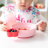 A toddler enjoys breakfast with from Bumkins Silicone Grip Bowl: Pink, with a matching cup and utensils, at a white table.