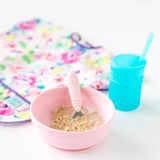 Bumkins Silicone Grip Bowl: Pink with cereal and milk, beside a blue cup with a straw and a colorful bib on a white background.